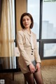 A woman in a beige suit standing in front of a window.